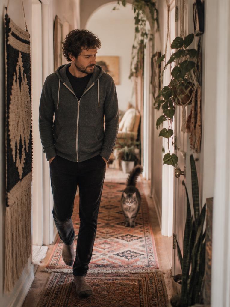 Man wearing the Juniper Slate Zip Hoodie walking a rug-lined boho hallway with a longhair cat